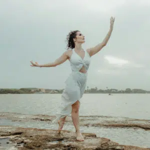 A woman stands on a beach, reaching up into the air to encompass the energy of Aquarius season.