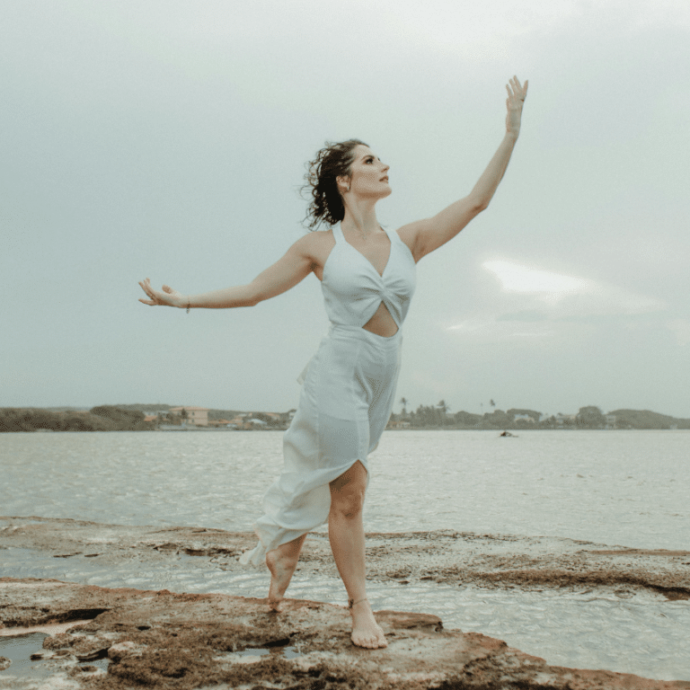 A woman stands on a beach, reaching up into the air to encompass the energy of Aquarius season.