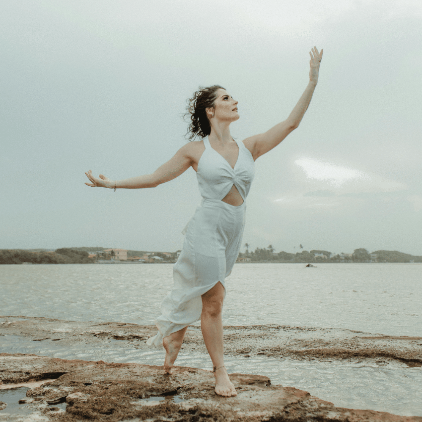 A woman stands on a beach, reaching up into the air to encompass the energy of Aquarius season.