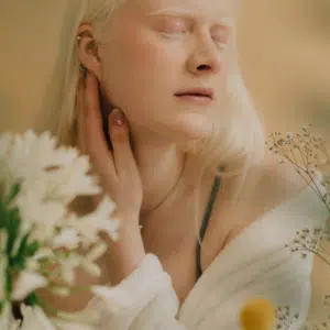 A woman with pale skin and hair has her eyes closed and is surrounded by daisies.