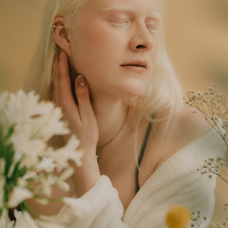 A woman with pale skin and hair has her eyes closed and is surrounded by daisies.