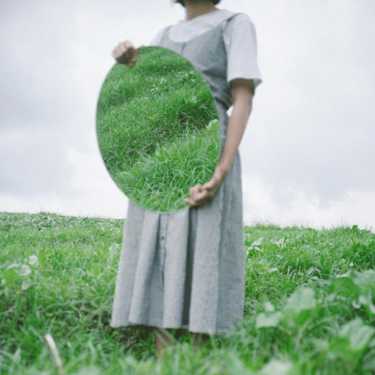 A woman holds a mirror in front of her, reflecting a field of green grass.