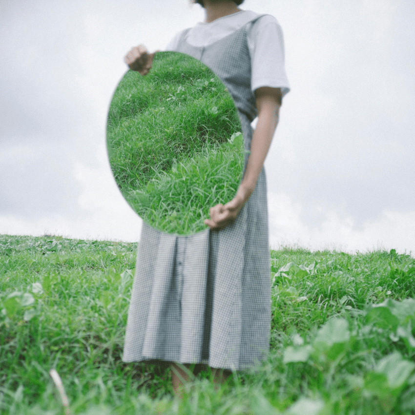 A woman holds a mirror in front of her, reflecting a field of green grass.