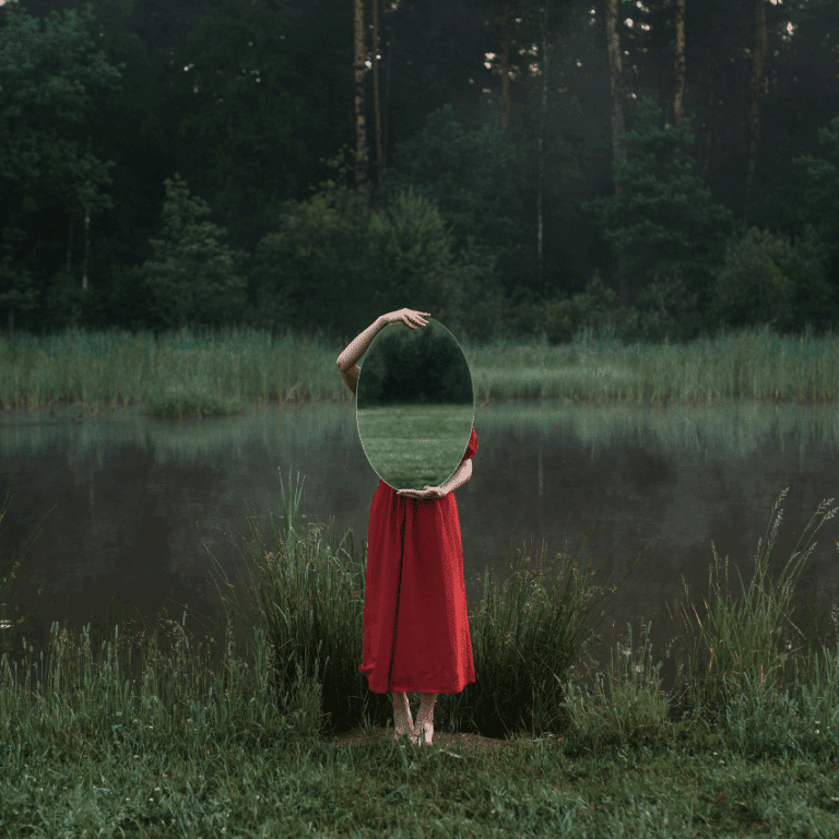 A person wearing a red dress stands in a green field and holds a mirror, reflecting the field.