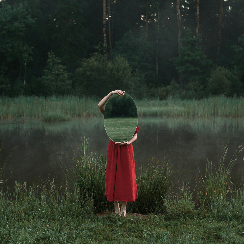 A person wearing a red dress stands in a green field and holds a mirror, reflecting the field.