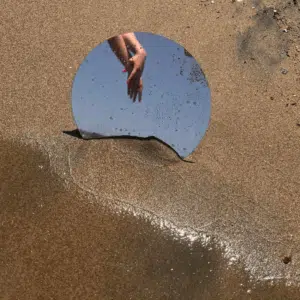 A mirror in the sand shows two hands grasping each other with a blue sky.