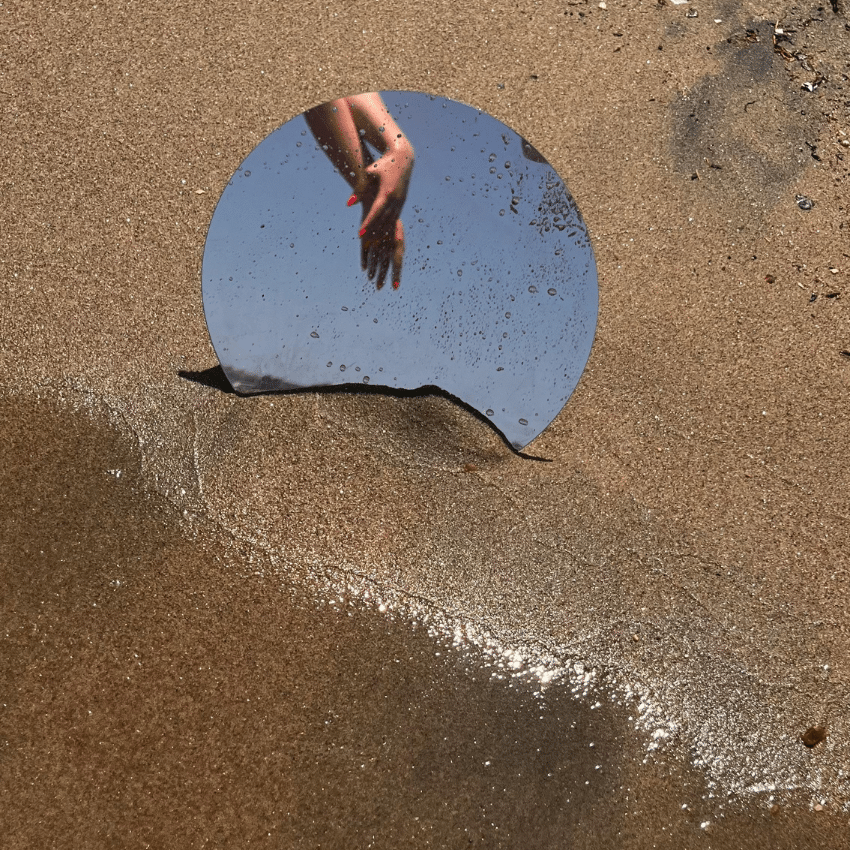 A mirror in the sand shows two hands grasping each other with a blue sky.