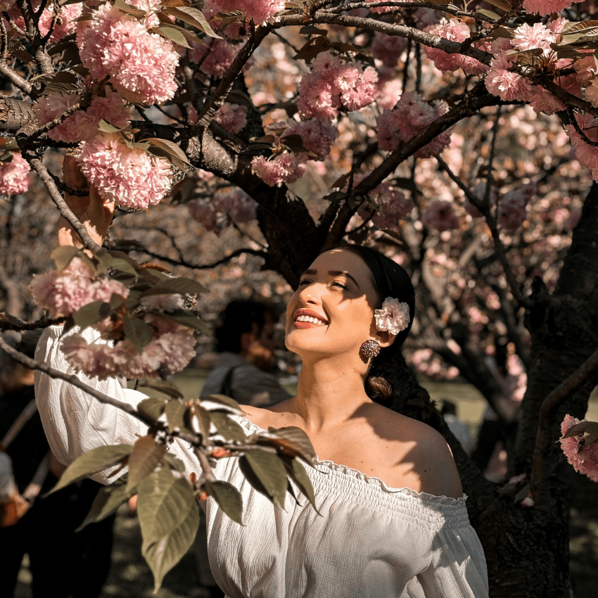 A woman in a white dress stands under a blossoming tree and smiles.