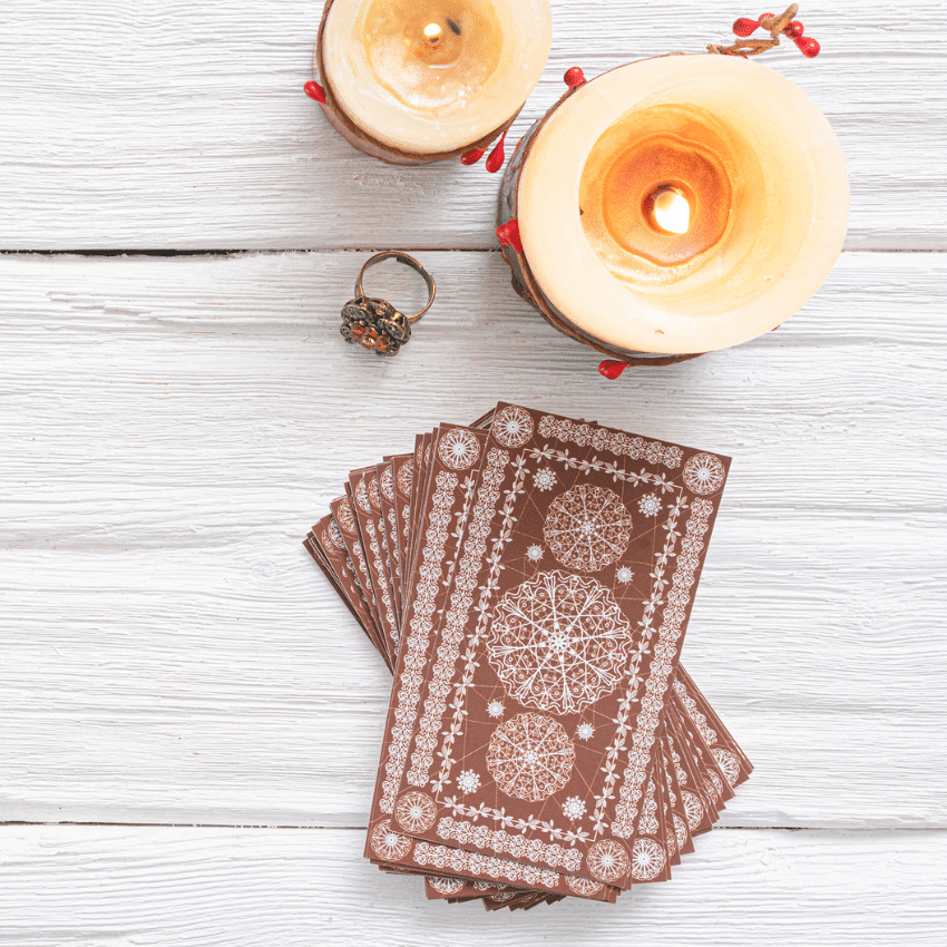 A stack of brown Tarot cards sits on a table next to 2 candles and a ring of colorful crystals.