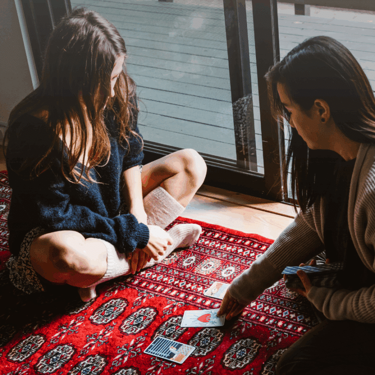 Two people sit on a rug with a stack of Tarot cards in between them.