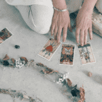 someone crouching on the ground and holding their hands over tarot cards with a line of different color crystals