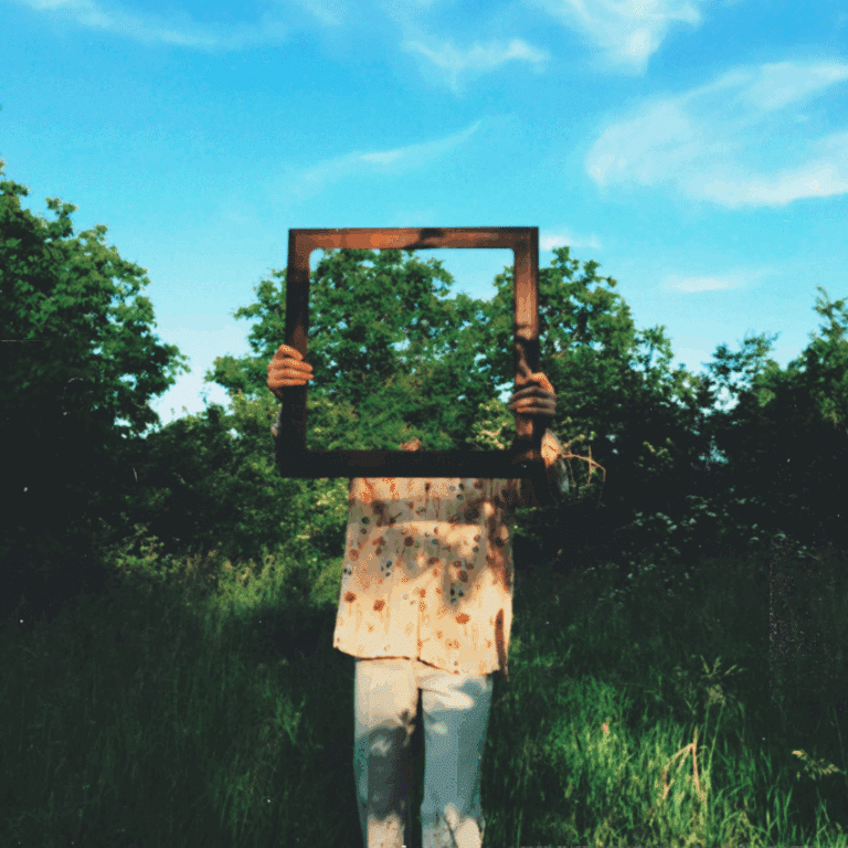 A person wearing white clothing holds up a mirror in front of their face, reflection the forest around them.