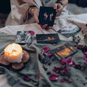 someone holds black Tarot cards in front of a table scattered with flower petals and a lit candle.