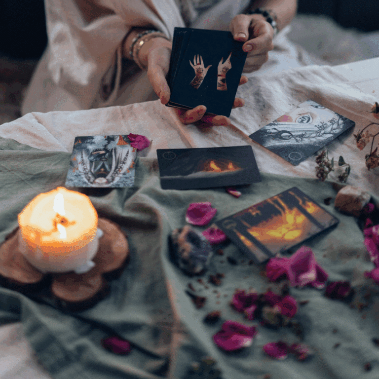 someone holds black Tarot cards in front of a table scattered with flower petals and a lit candle.