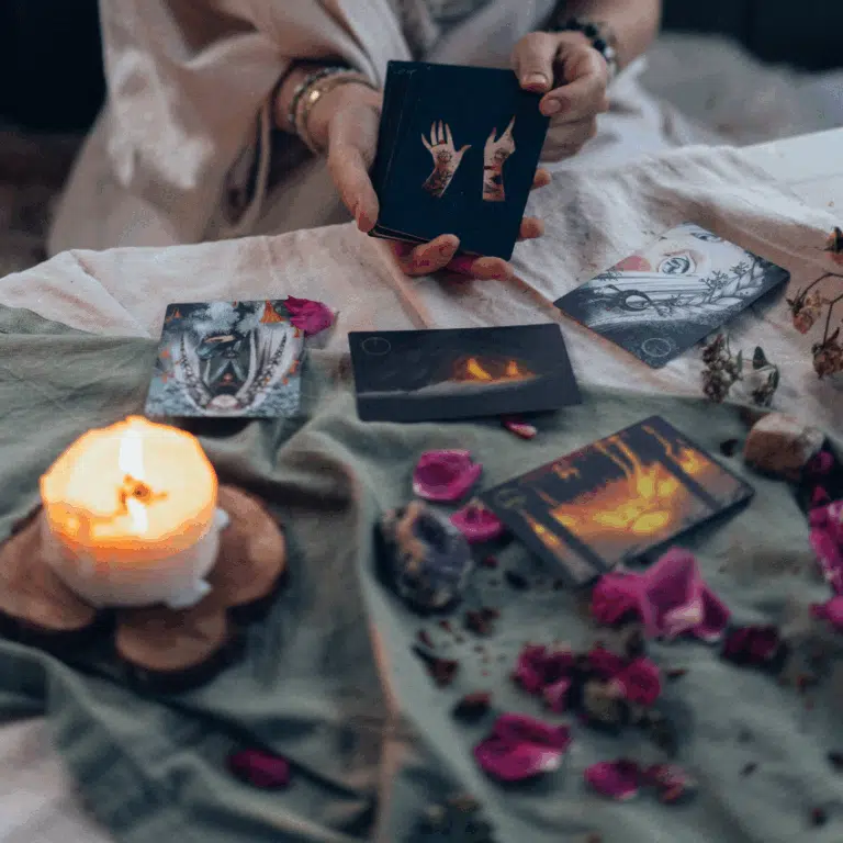 someone holds black Tarot cards in front of a table scattered with flower petals and a lit candle.