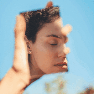 A woman holds up a mirror reflecting her face. There is a blue sky and blurred trees in the background.