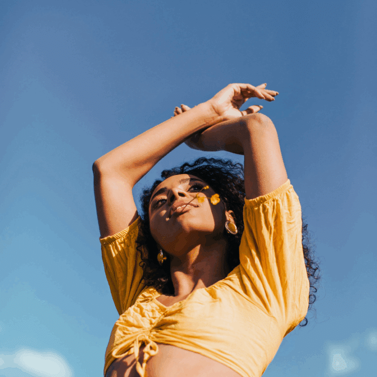 A woman of color with long black hair holds her hands in the air above her head. She is wearing a bright yellow 2 piece clothing set. The blue sky in the background sets the stage for abundance.