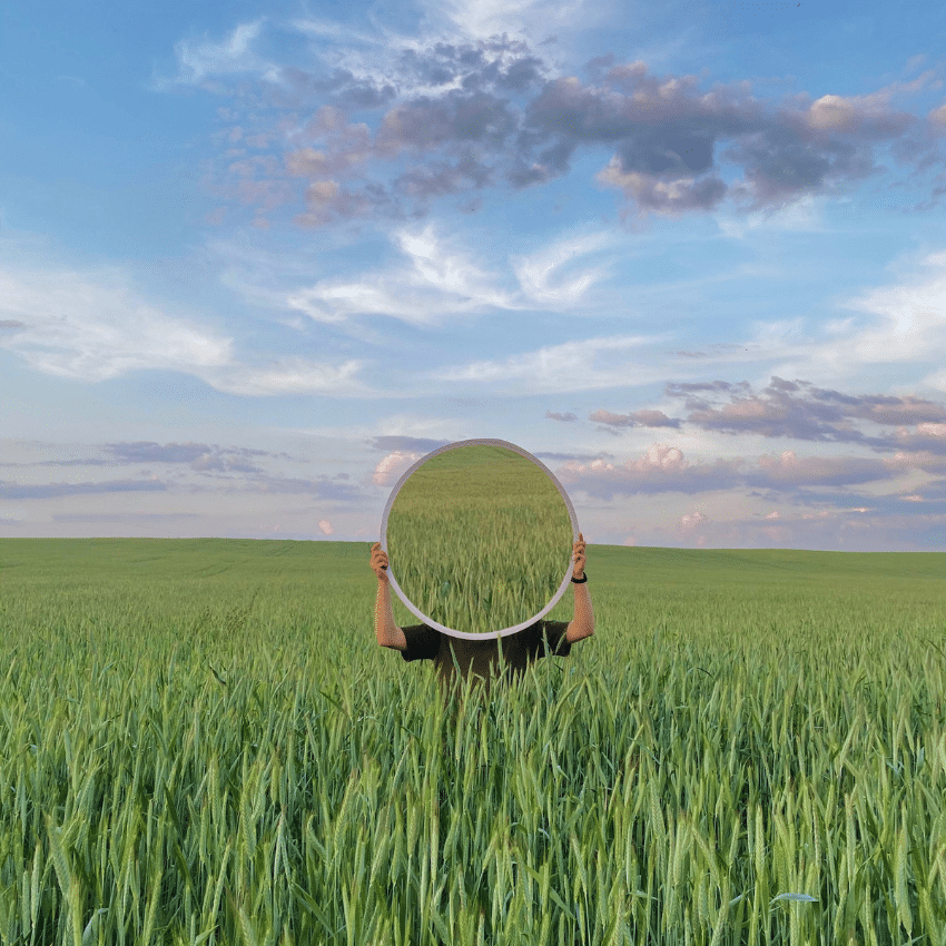 A person holds up a circular mirror in a field, reflecting a field of tall, green grass back.