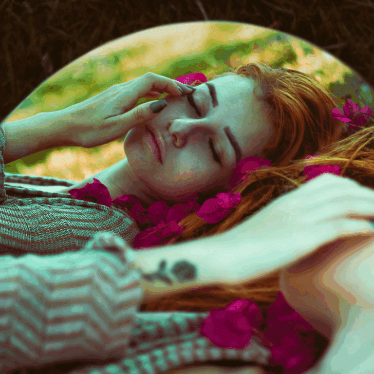 A woman with bright red hair lays with some pink flower petals next to a mirror, reflecting her peaceful expression back.