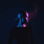 A woman wearing black with her head turned to the side is illuminated in dark blue and purple light.