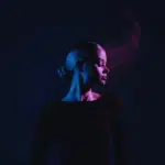 A woman wearing black with her head turned to the side is illuminated in dark blue and purple light.