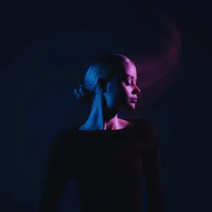 A woman wearing black with her head turned to the side is illuminated in dark blue and purple light.