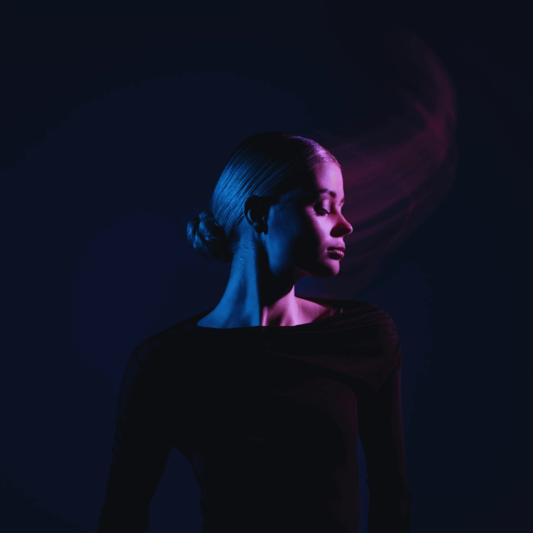 A woman wearing black with her head turned to the side is illuminated in dark blue and purple light.