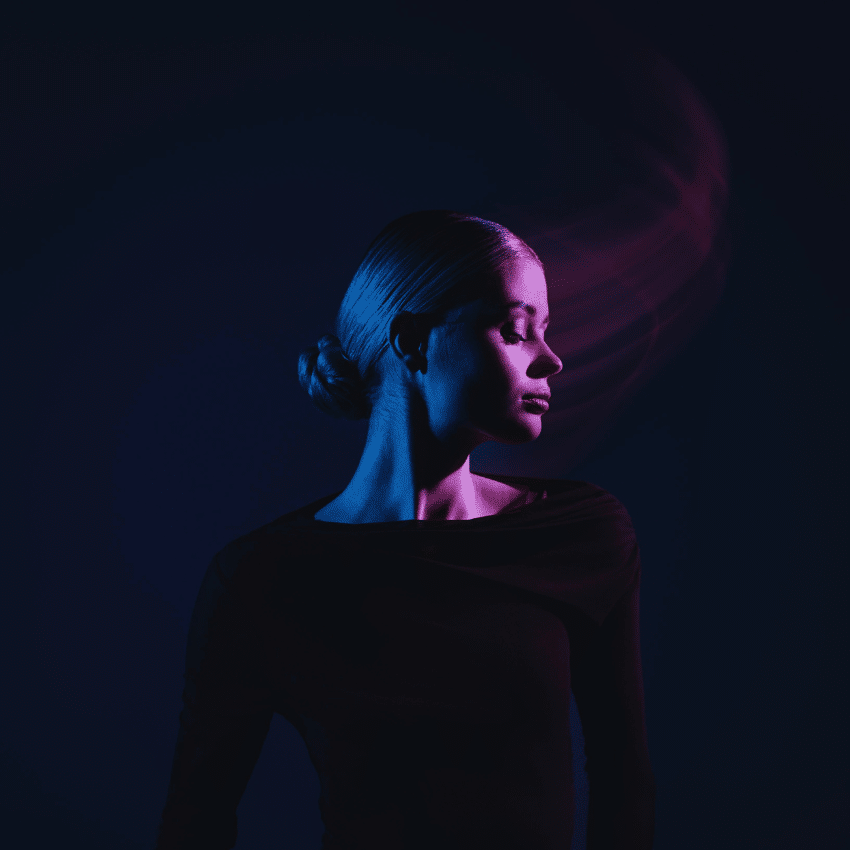 A woman wearing black with her head turned to the side is illuminated in dark blue and purple light.