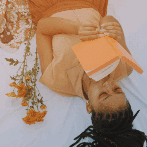 A woman lays next to a bouquet of orange flowers and holds an orange book above her face.