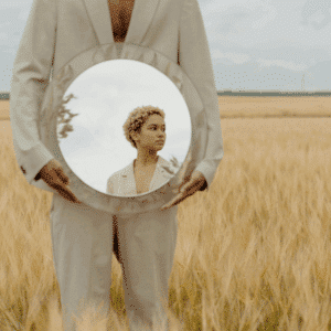 A person wearing a cream-colored suit stands in a field of wheat holding a mirror reflecting a person and the sky.