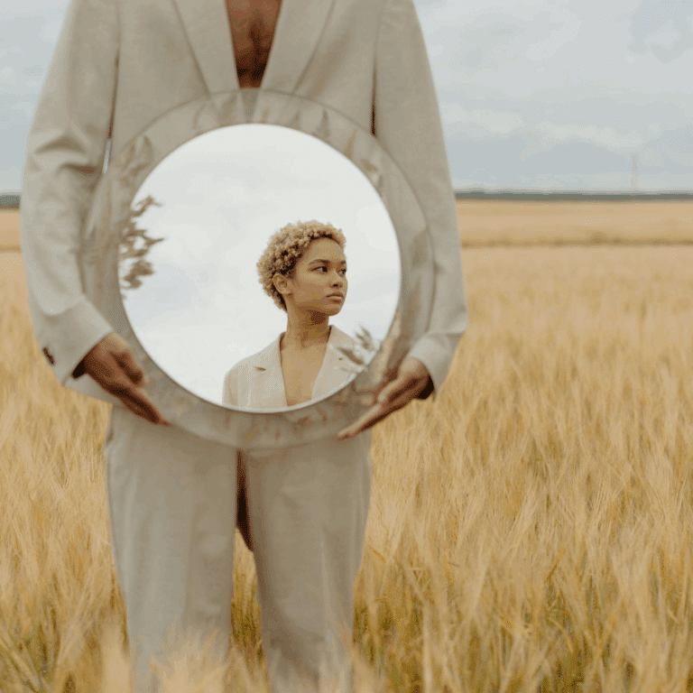 A person wearing a cream-colored suit stands in a field of wheat holding a mirror reflecting a person and the sky.