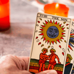 A hand holds a handful of Tarot cards with The Sun card in the front. Two orange candles burn in the background.