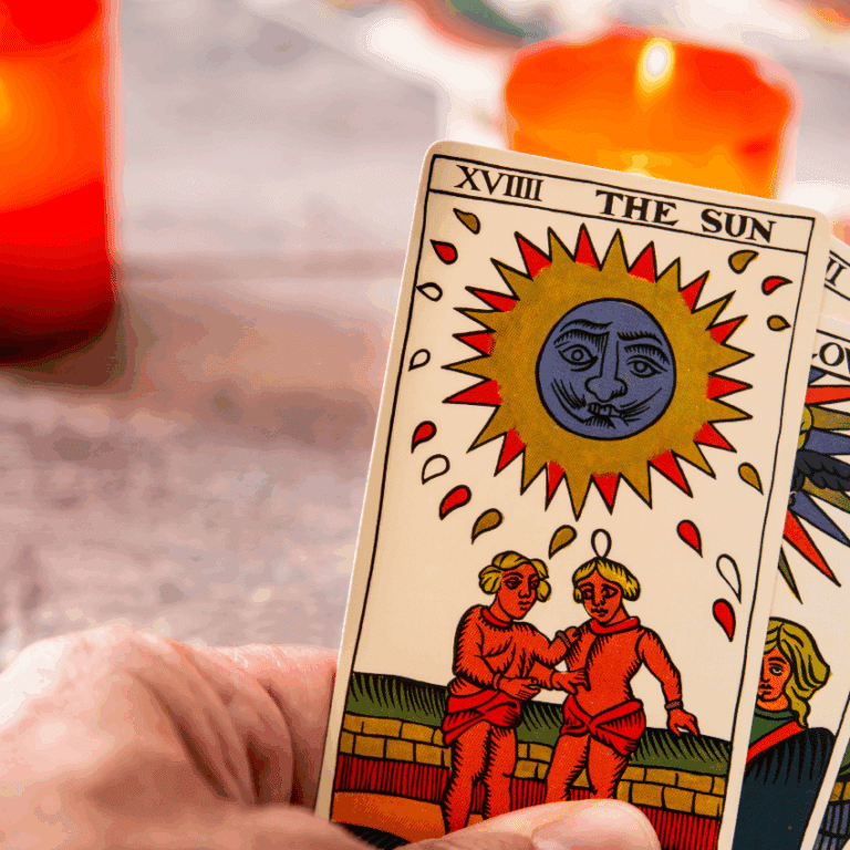 A hand holds a handful of Tarot cards with The Sun card in the front. Two orange candles burn in the background.