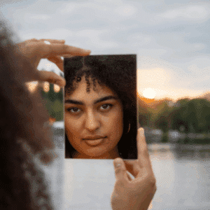 A woman with dark hair and eyes looks into a square mirror, reflecting her face back the the viewer. The background is a lake with trees at sunset.