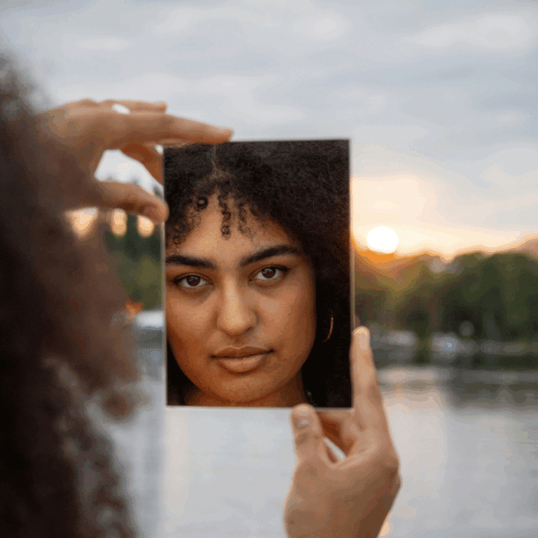 A woman with dark hair and eyes looks into a square mirror, reflecting her face back the the viewer. The background is a lake with trees at sunset.