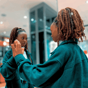A black woman reaches up to her reflection in a mirror.