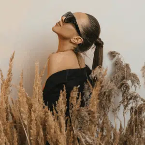 A brunette woman with a ponytail and sunglasses leans her head back amongst stalks of wheat, representing Virgo season in August.