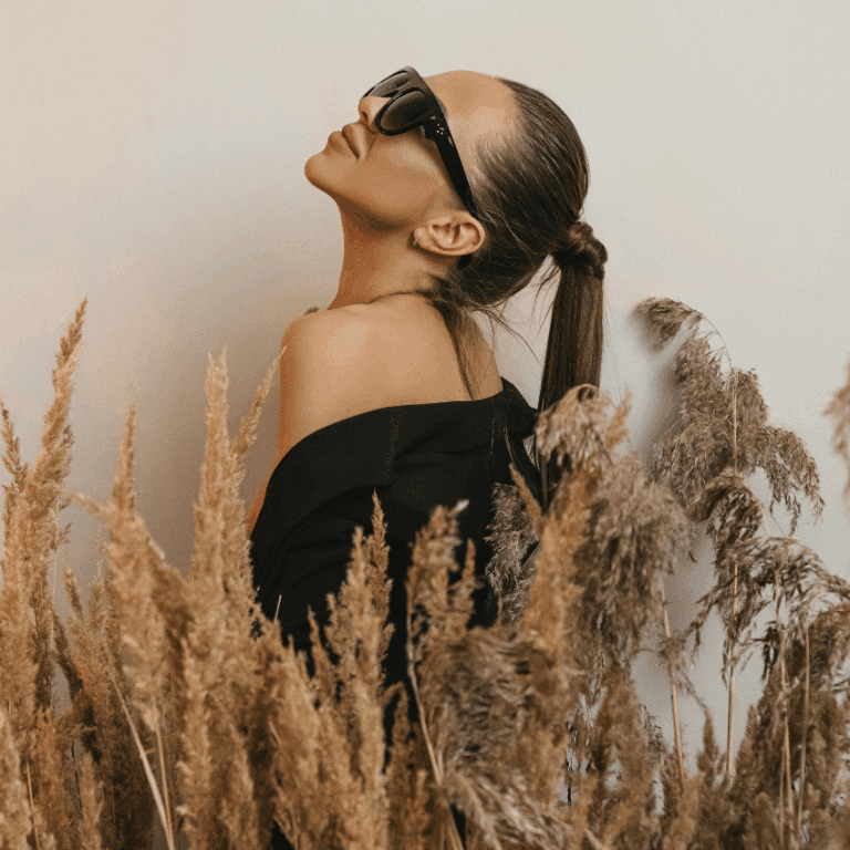A brunette woman with a ponytail and sunglasses leans her head back amongst stalks of wheat, representing Virgo season in August.