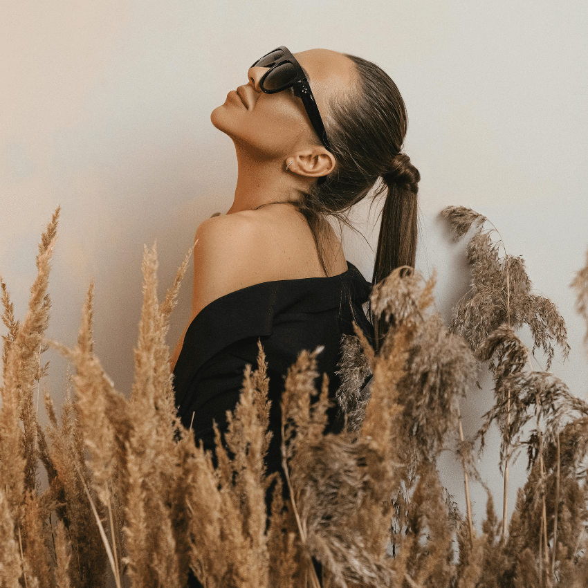 A brunette woman with a ponytail and sunglasses leans her head back amongst stalks of wheat, representing Virgo season in August.