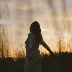 A woman, shadowed by the setting sun, walks through a field of tall grasses.