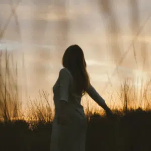 A woman, shadowed by the setting sun, walks through a field of tall grasses.