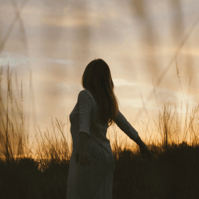 A woman, shadowed by the setting sun, walks through a field of tall grasses.