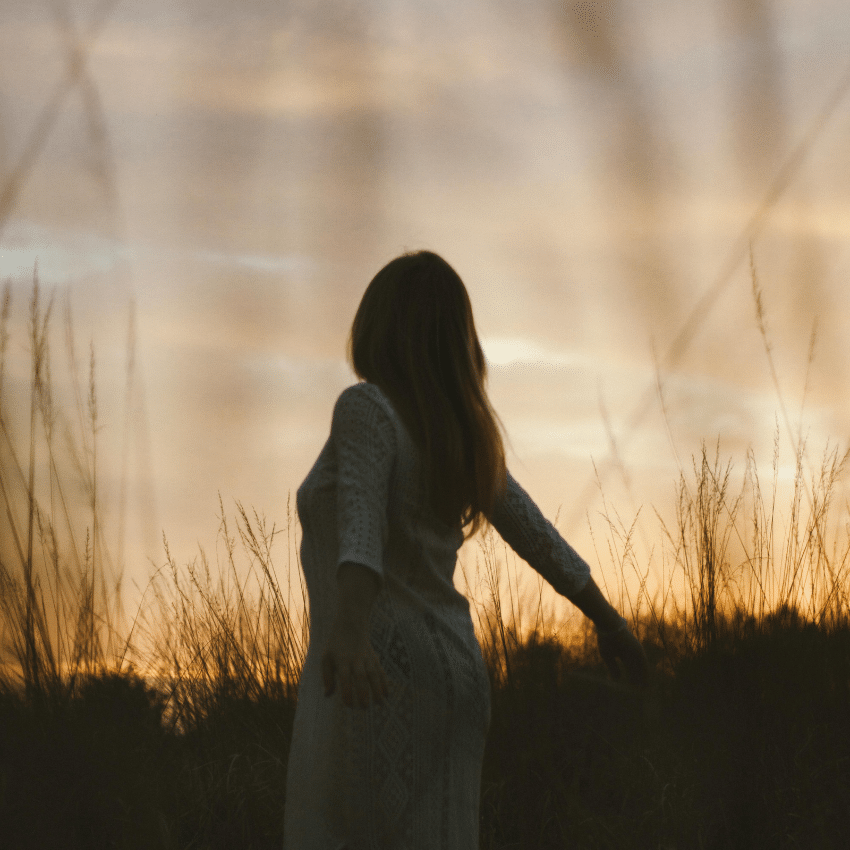 A woman, shadowed by the setting sun, walks through a field of tall grasses.