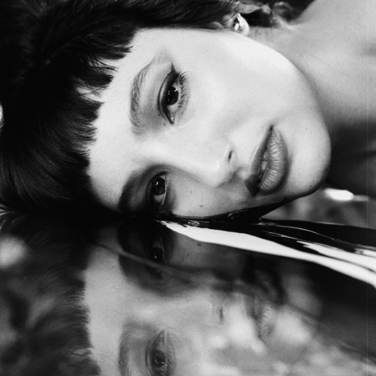 A black and white image of a woman laying on a mirror, her reflection showing up under her.