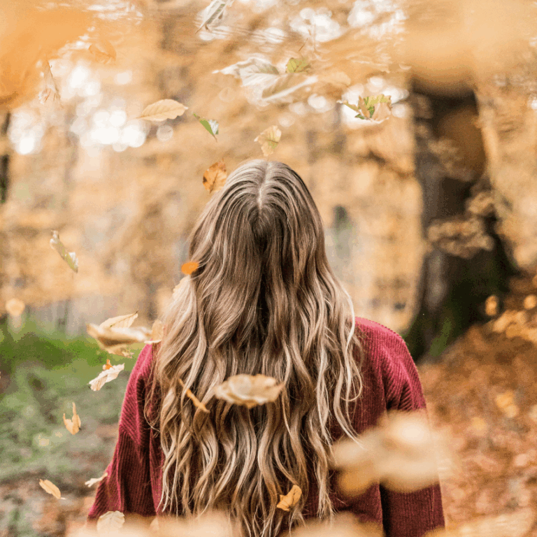 A woman faces a forest of trees with orange leaves, falling around her.