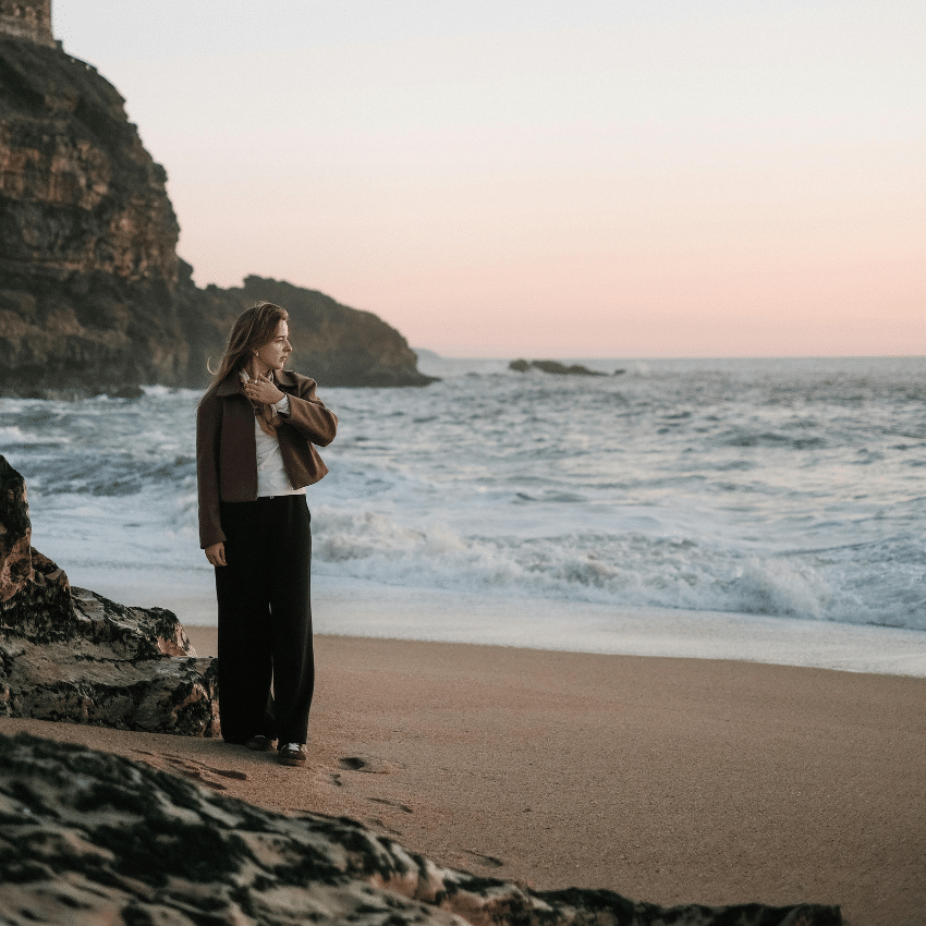 A woman stands on a rocky beach, peering into the water.