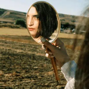 A woman looks into a handheld mirror, her reflection looking back at the viewer.