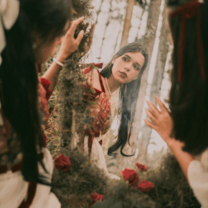 A woman wearing a gown looks in a mirror, her contemplative reflection shown to the viewer.