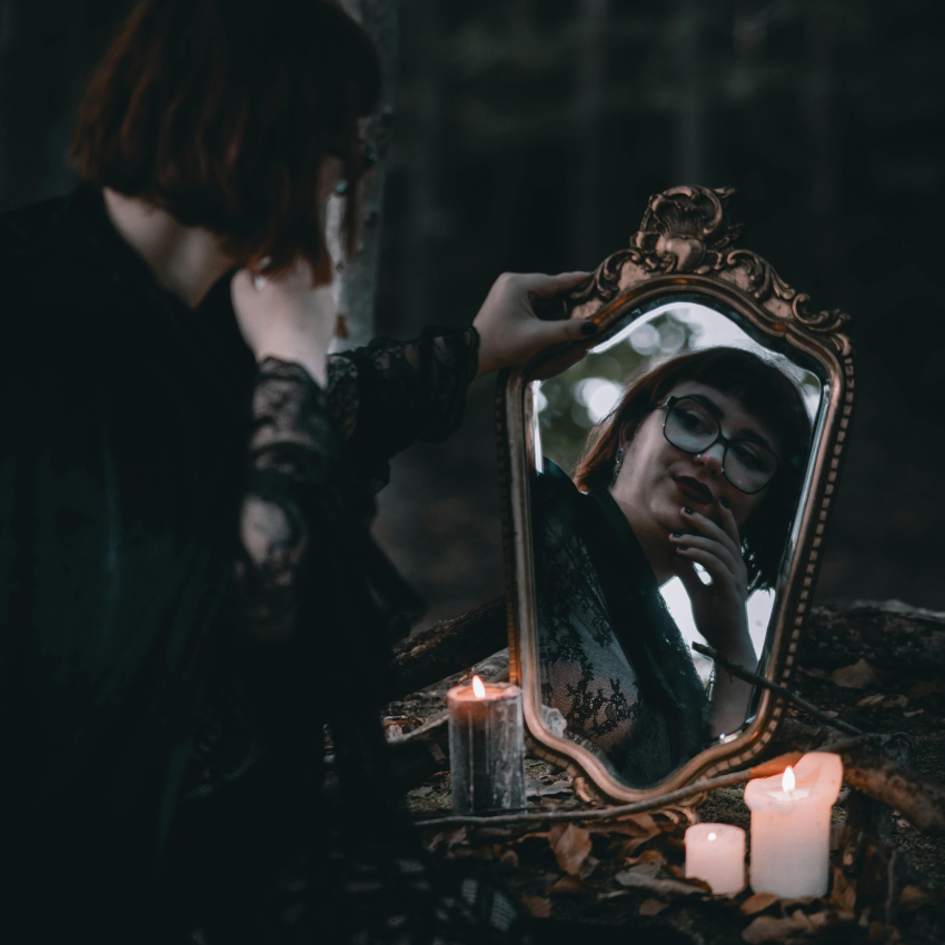 A dark night with a woman in a gold mirror, her reflection showing her face. The mirror is surrounded by candles.