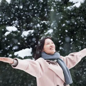 A woman wearing a pink jacket and a grey scarf holds her arms out embracing the falling snow to represent the astrological energy of December.